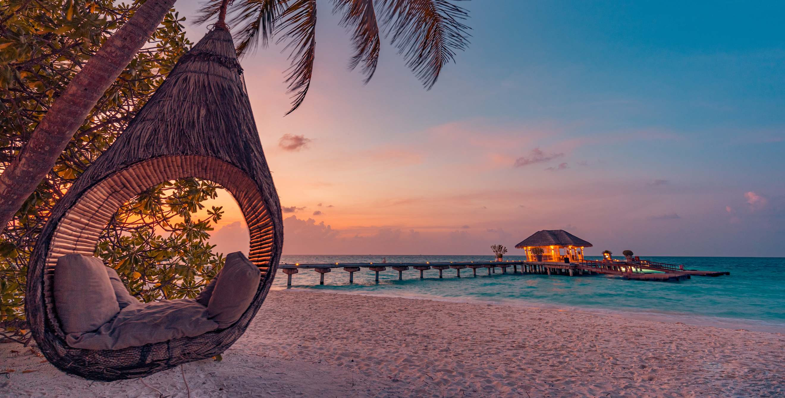 A seating pod hangs from a palm tree at a tropical beach resort at sunset.