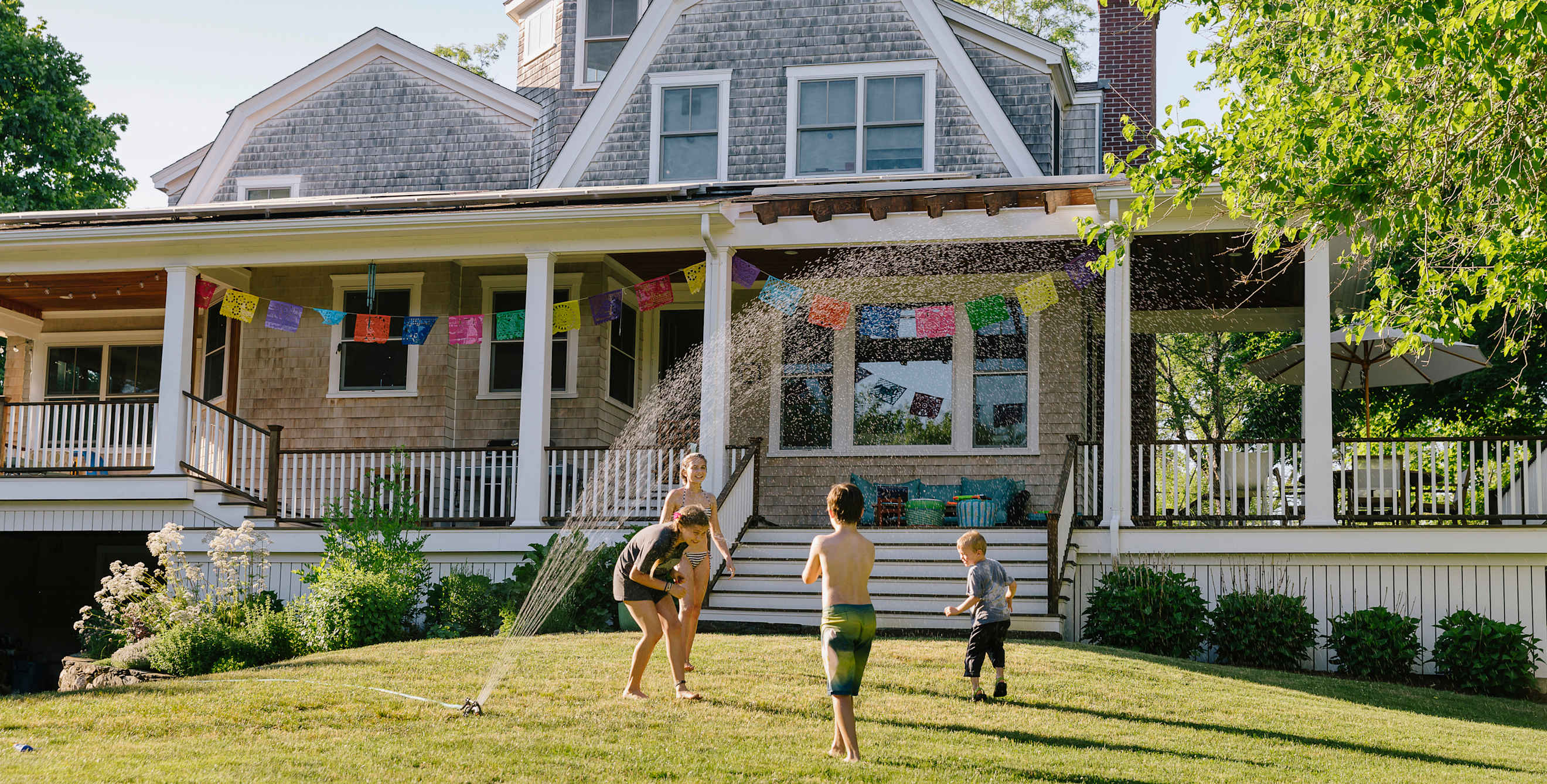 Kids play in a sprinkler in front of a wood-shingled house.