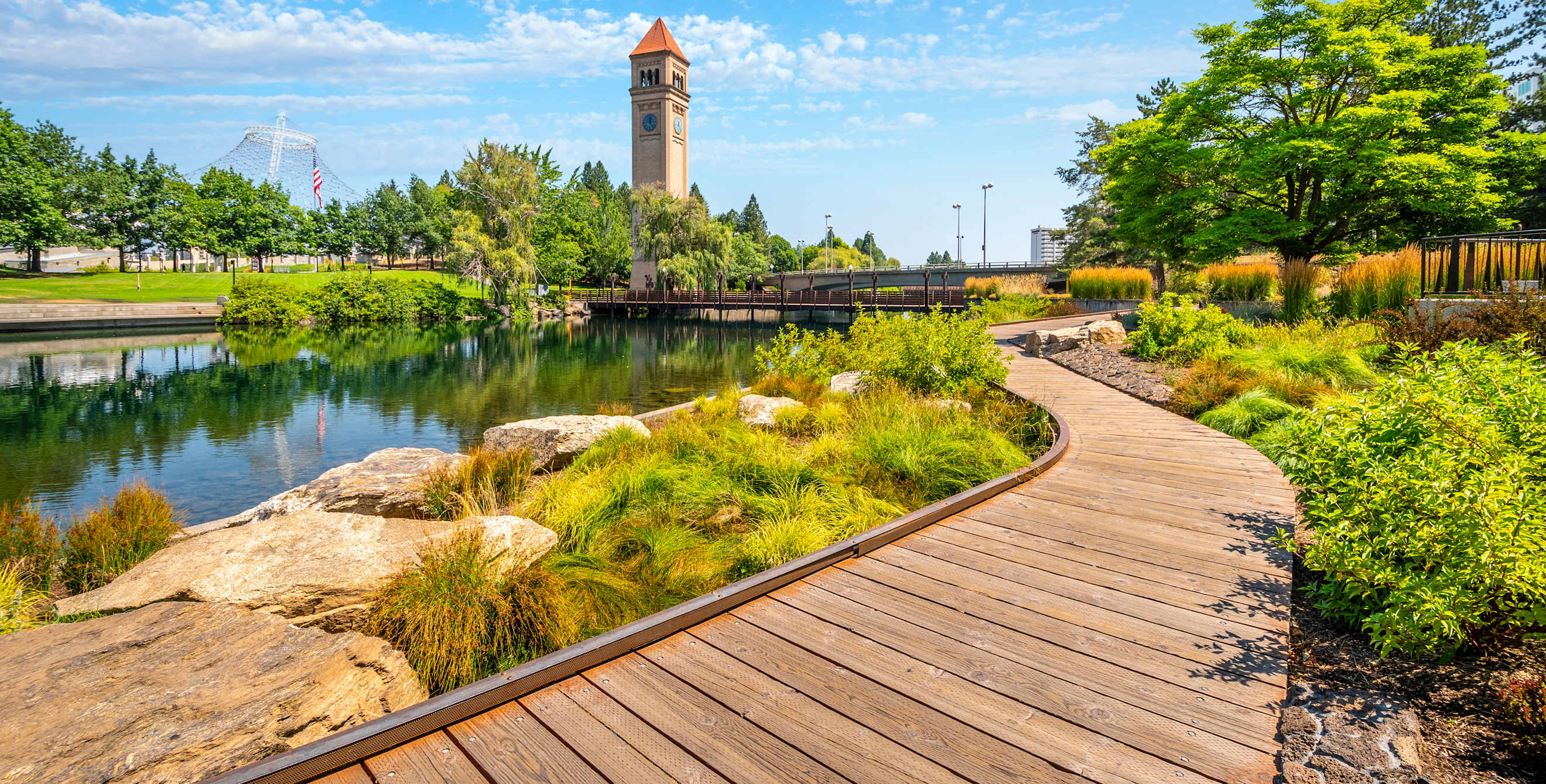 A boardwalk path beside a river with a clock tower in the distance.