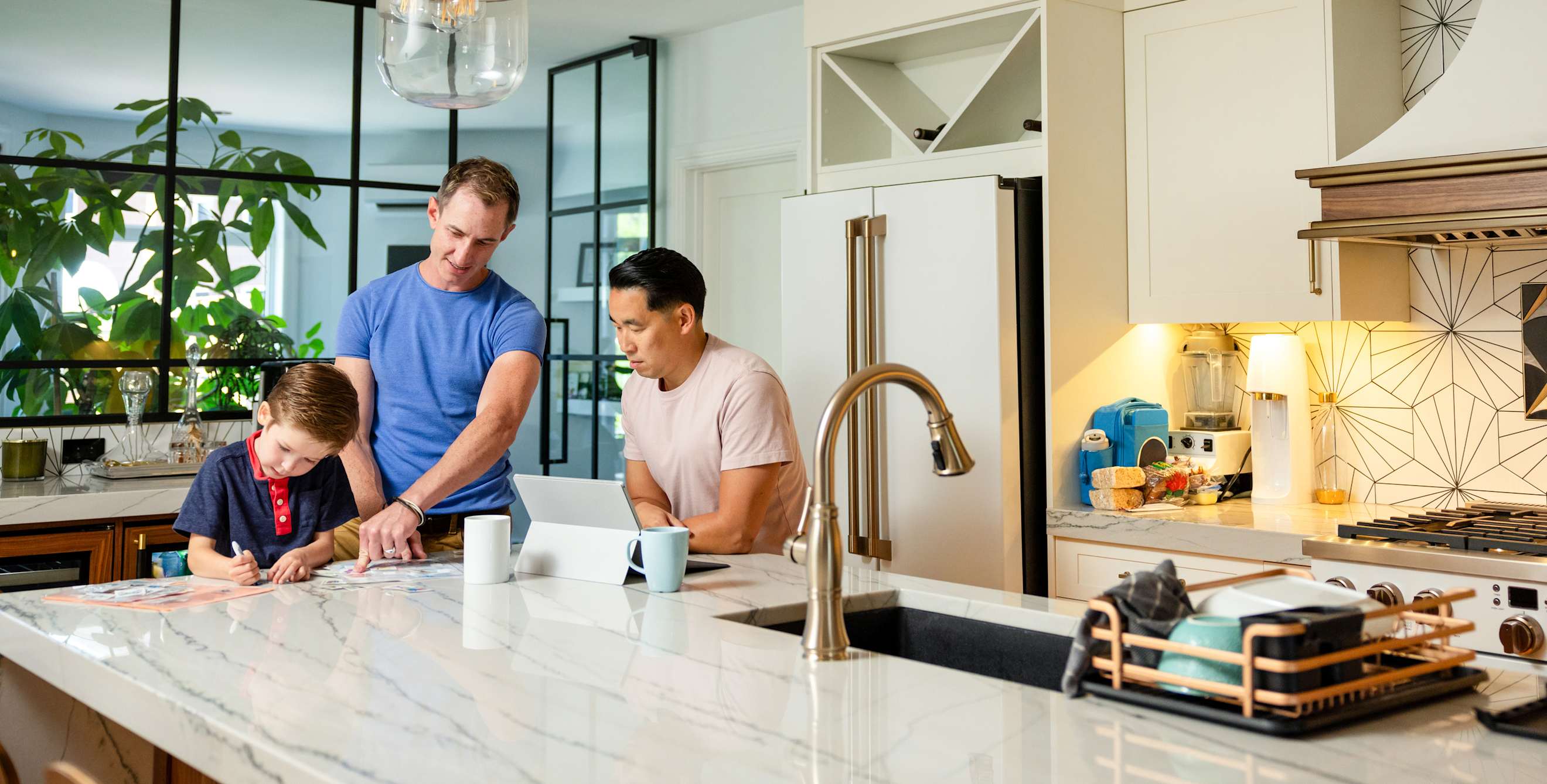 Two dads stand beside their young son at a modern kitchen island.