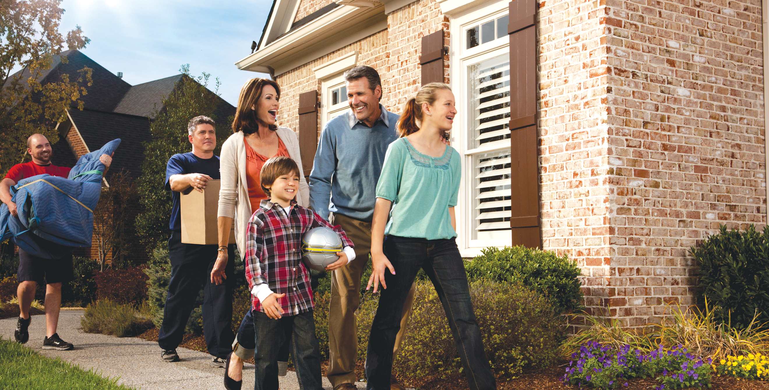 A family of four walk toward their house with a pair of movers behind them.