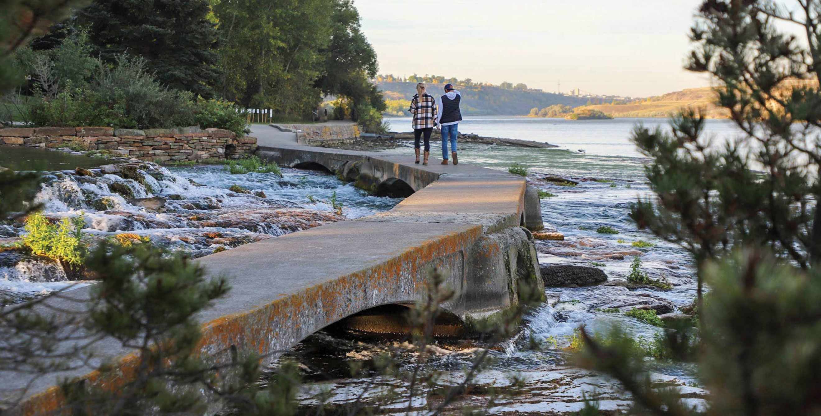 people walk along a scenic pathway near trees and water in Great Falls, Montana