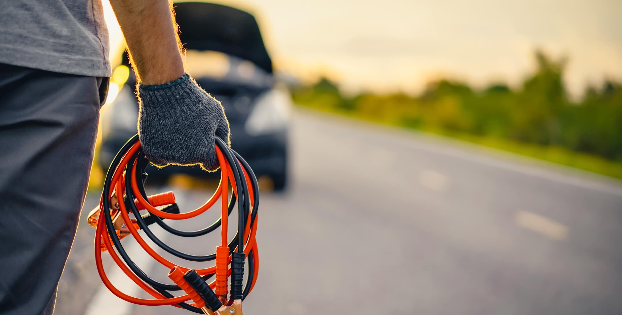 A driver walks toward their car with the hood up with jumper cables.