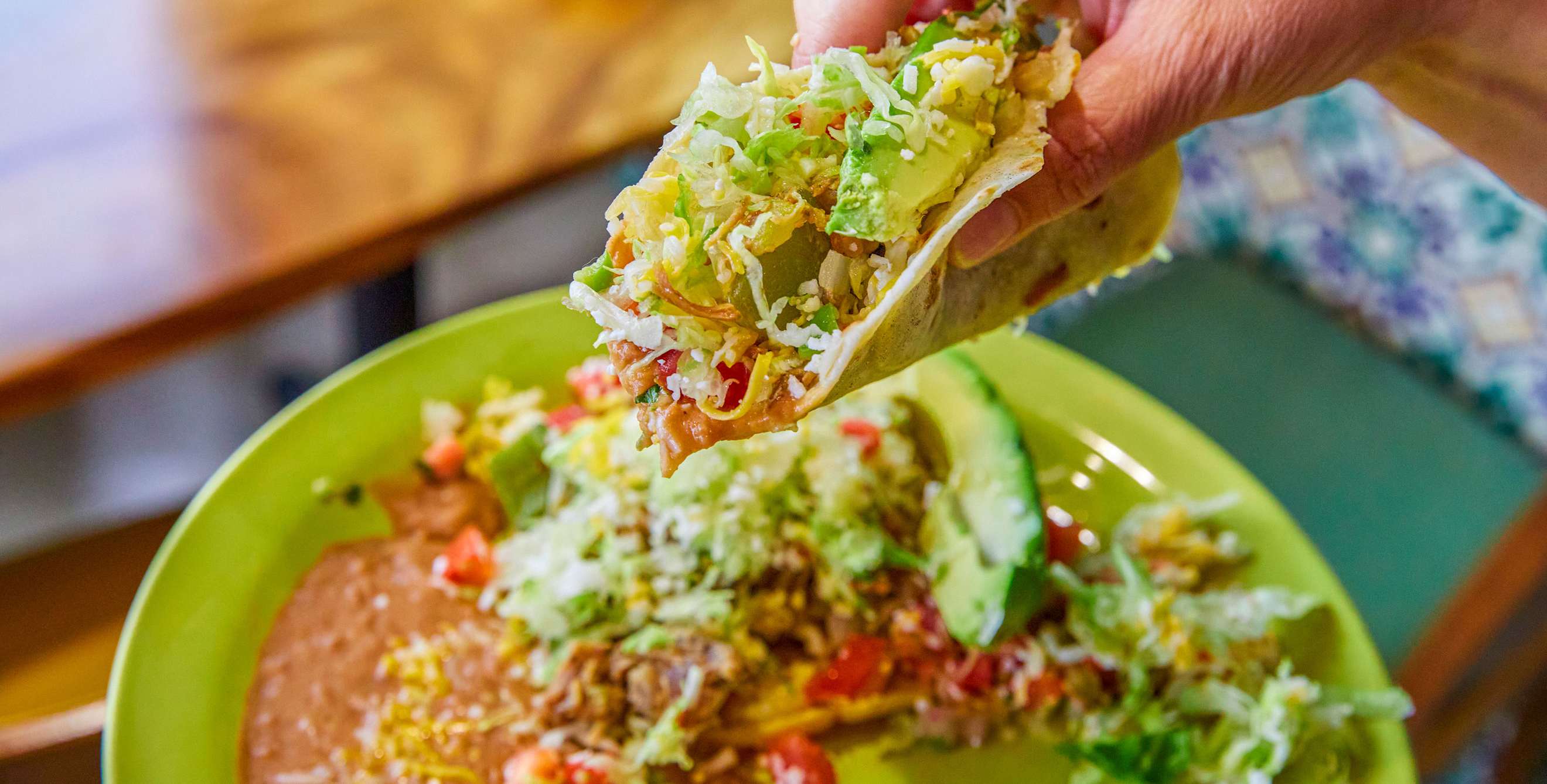 A hand holds an overstuffed soft taco over a platter of refried beans and salad.