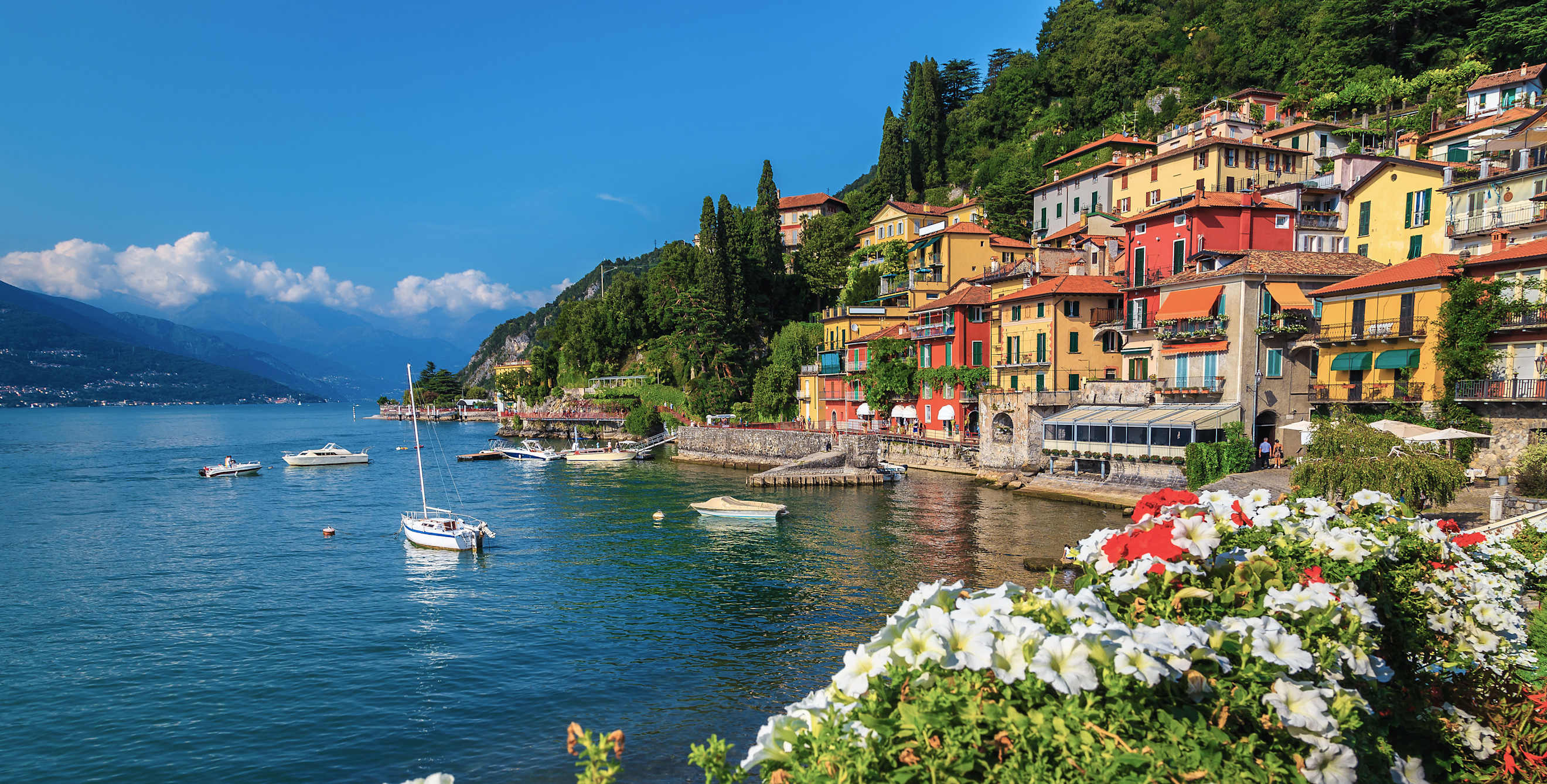 Flowery fence and colorful mediterranean buildings on the waterfront. Anchored boats in the harbor of Varenna, lake Como, Lombardy, Italy