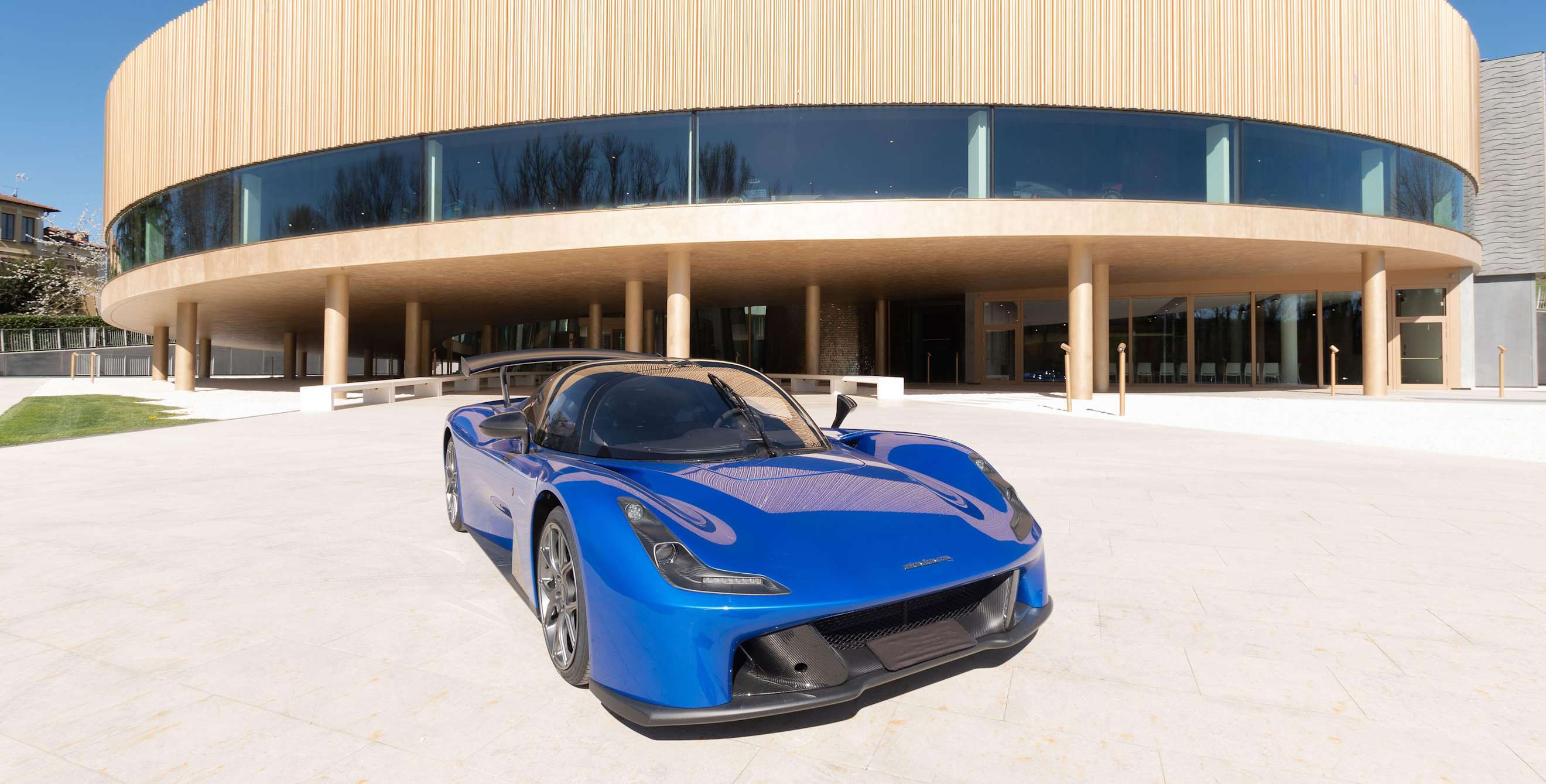 A blue Dallara Stradale sports car on a plaza in front of a curved, modern building