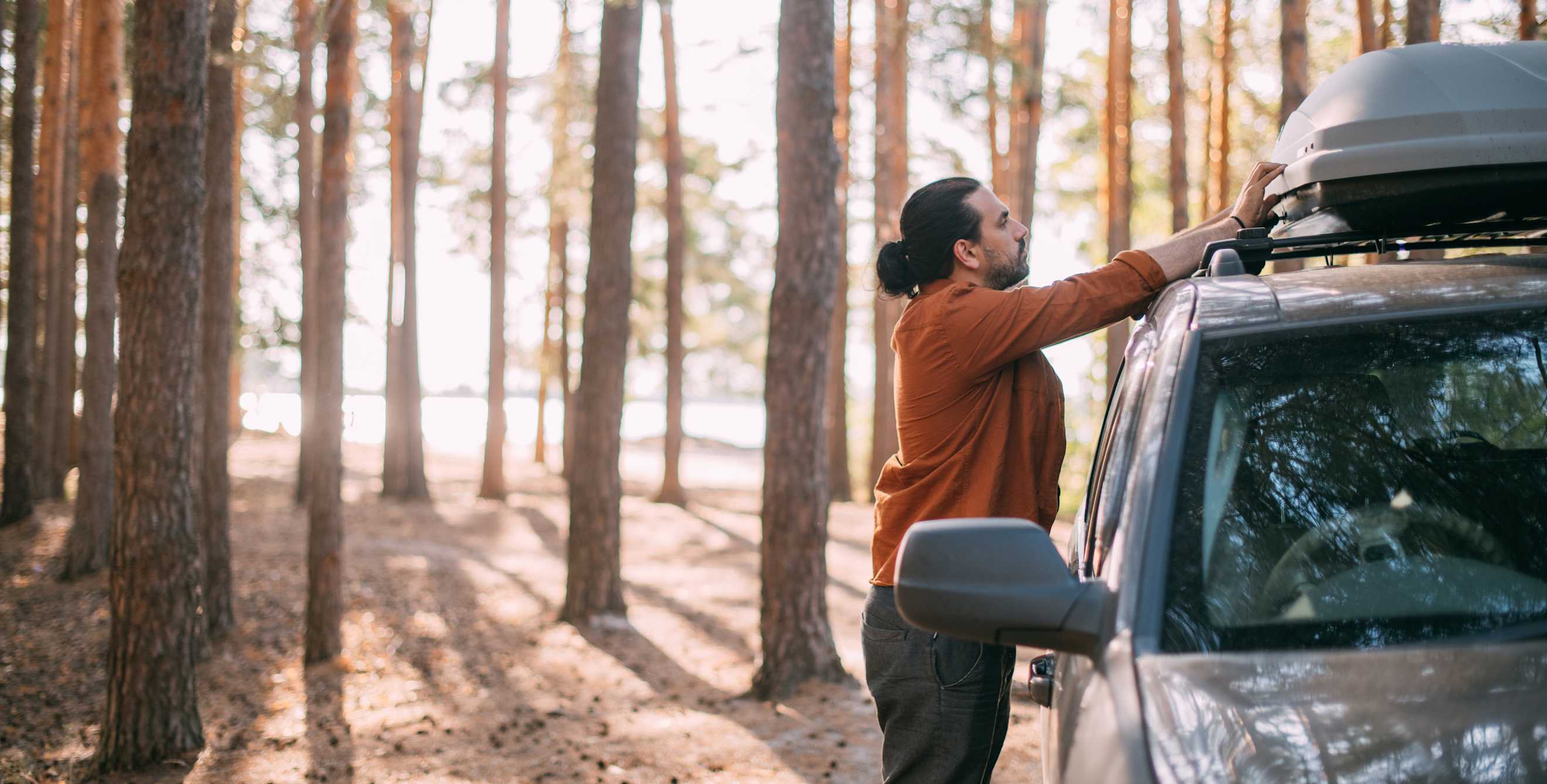 A person opens a roof-top storage box on their SUV parked in forest.