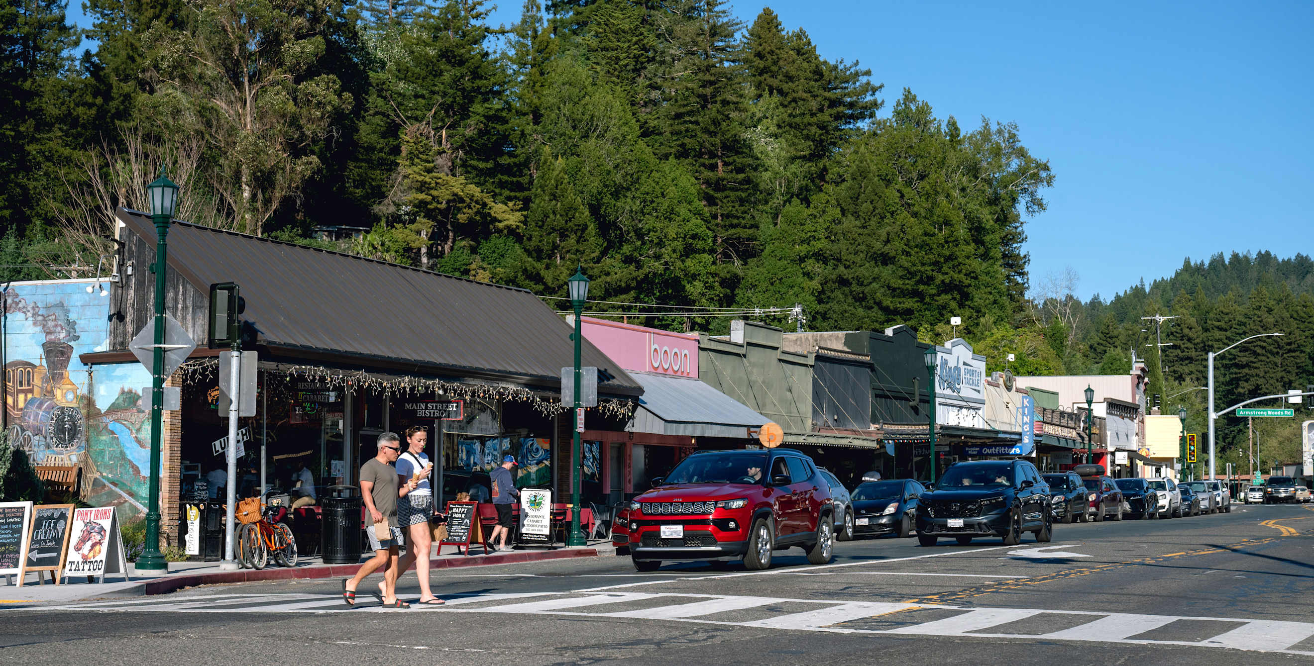 people walk across the street in a picturesque small town with trees and mountains visible against a blue sky