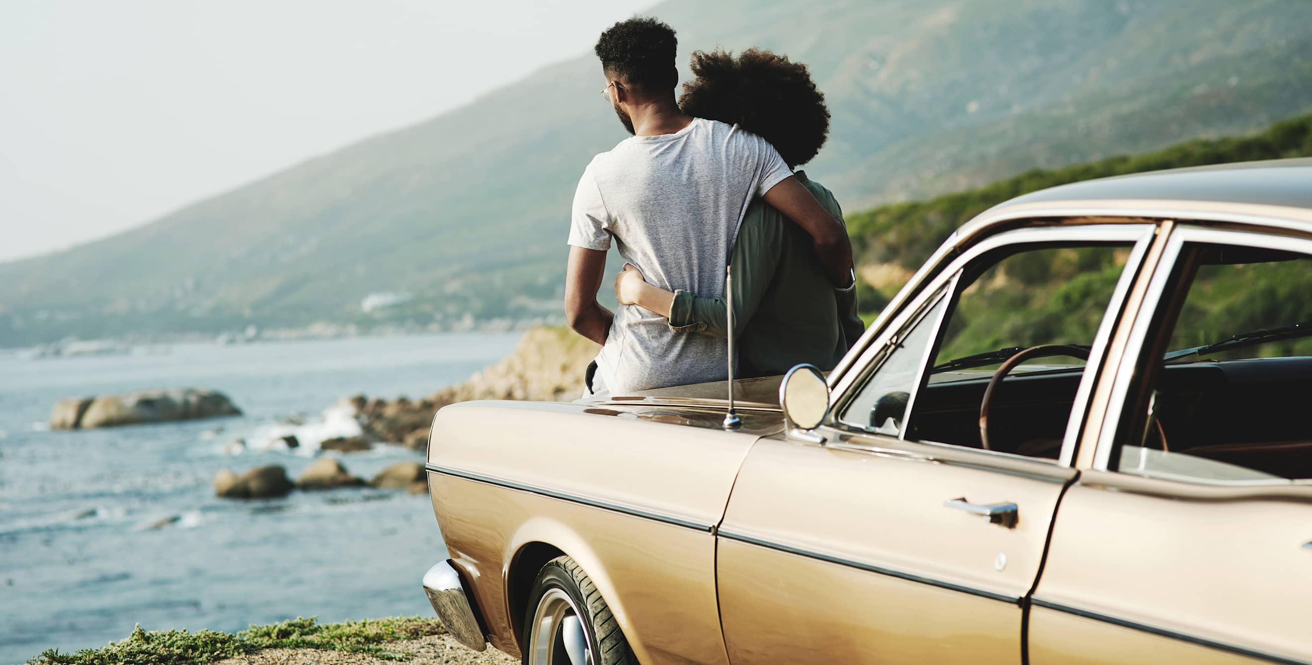 A couple lean against the hood of their gold vintage car.