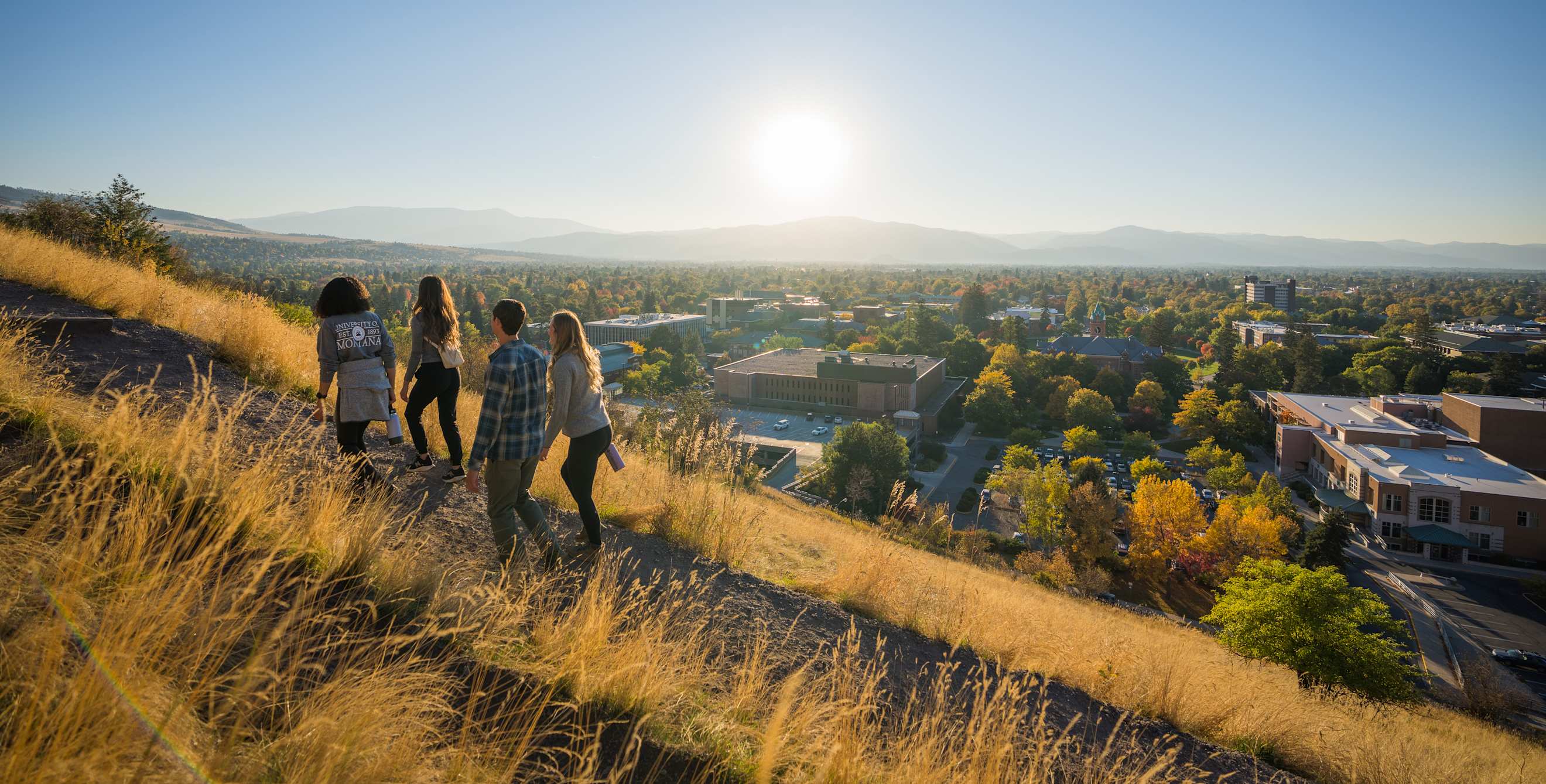 four people walk up a mountain with dry grass and a view of Missoula, Montana