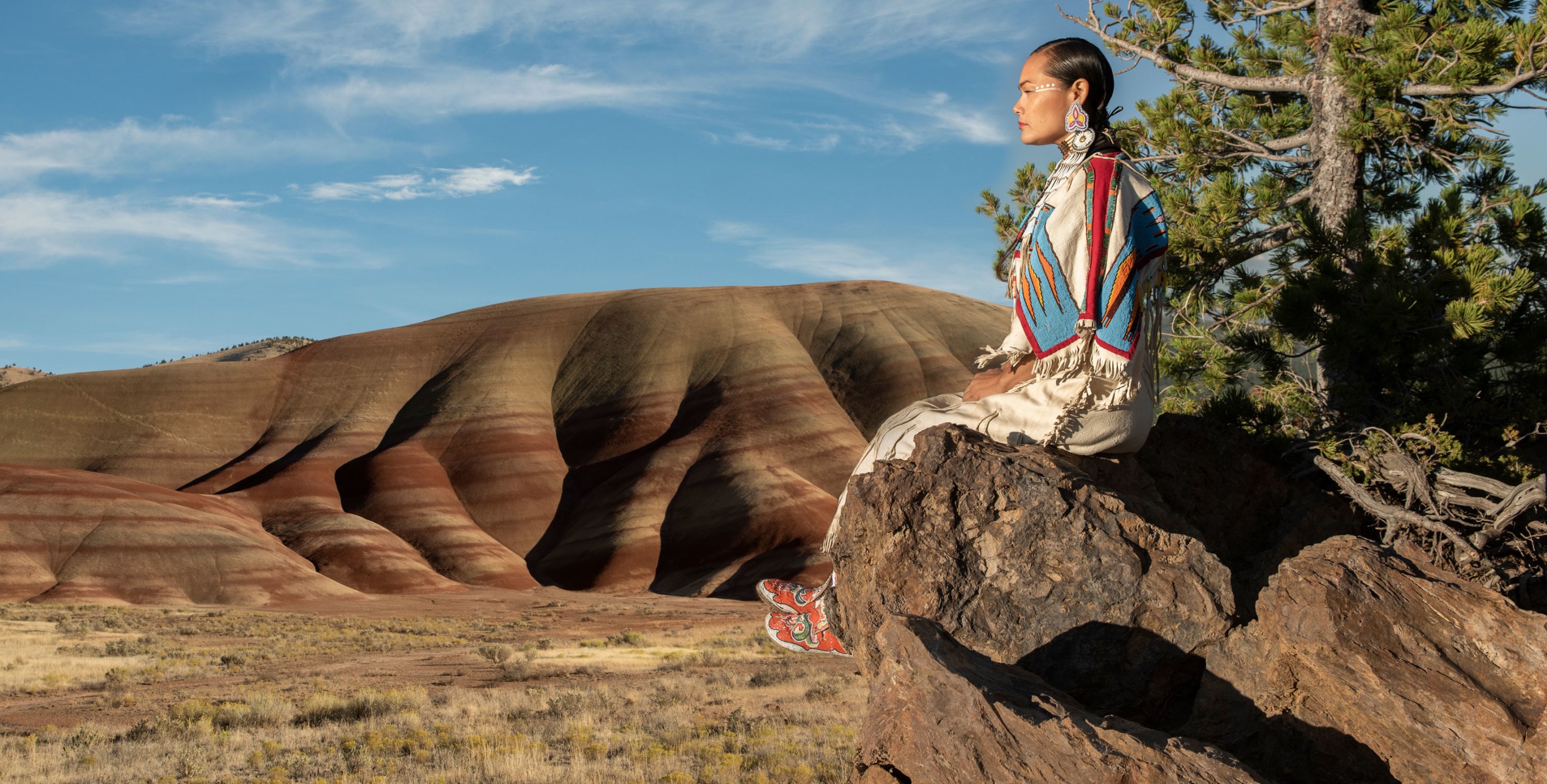 Acosia M. Red Elk in a buckskin dress sitting on a rock overlooking the Oregon landscape.