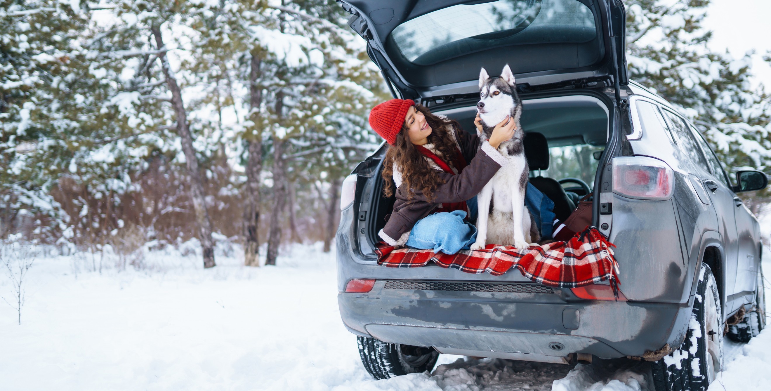 A woman sits in the back of her SUV with her husky dog on a snowy day.