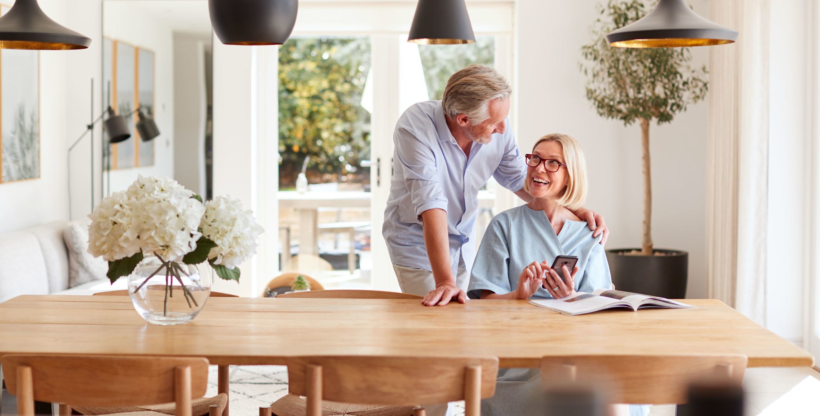 two smiling older adults at a wooden table looking through a catalog; the woman is holding her phone