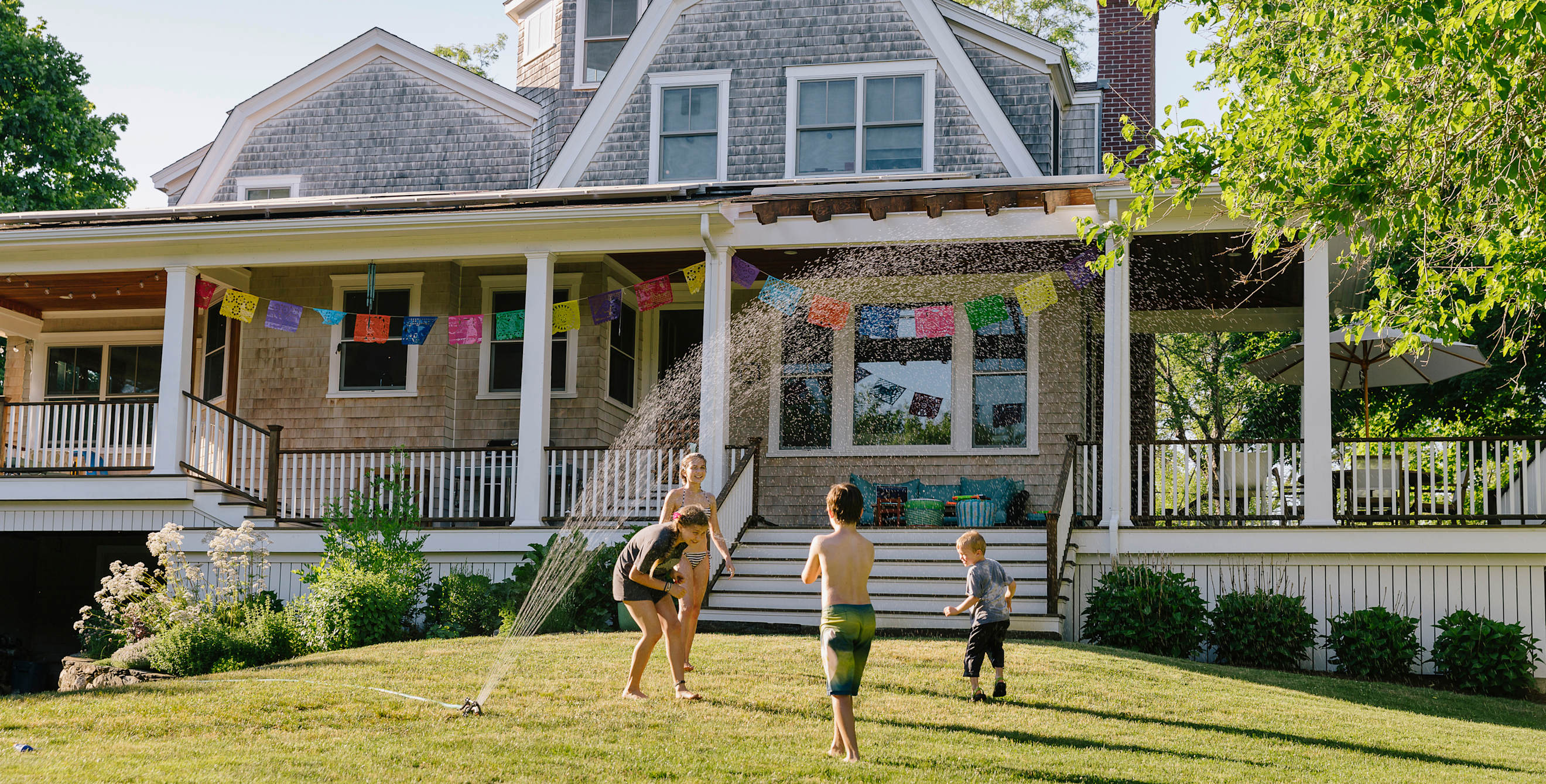 Kids play in a sprinkler in front of a wood-shingled house.