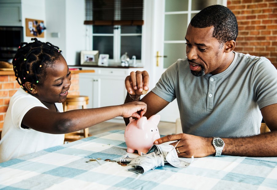 Dad and daughter put money into a piggy bank at the dinner table, image