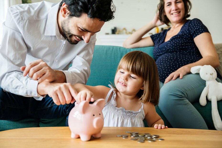 father helps daughter put coins into a piggy bank with mom in background, image