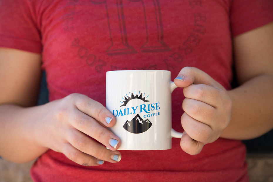 two hands hold a white mug with the Daily Rise coffee shop logo on it.