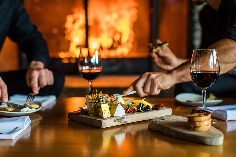 cheese and charcuterie board with glass of red wine and cozy background fire at Juniper Bistro in Banff, Alberta