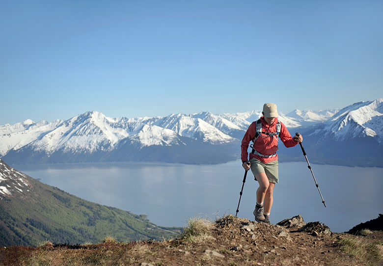 hiker in shorts uses trekking poles on Bird Ridge Trail above Turnagain Arm and snow-capped Chugach Mountains behind in Chugach State Park near Anchorage, Alaska, picture