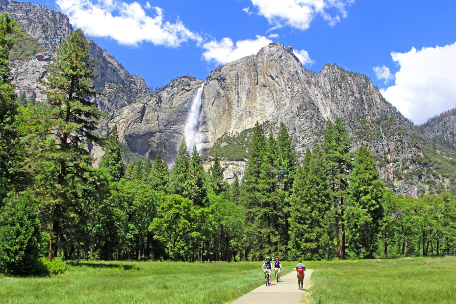Cyclists ride through Yosemite Valley with views of a waterfall in the background, image