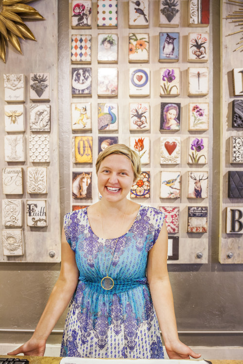 smiling woman greets warmly with display of hand-painted tiles in background at Passionflower Design, an eclectic gift store in Eugene, Oregon, picture