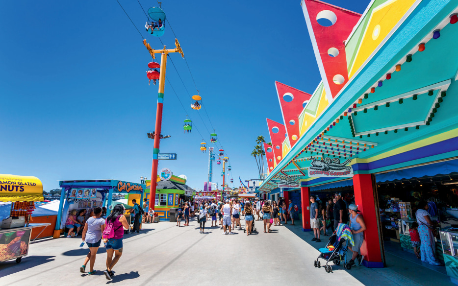 boardwalk visitors stroll past the colorful facades of food vendors and arcades as gondola riders pass from above at the Santa Cruz Beach Boardwalk, California, picture