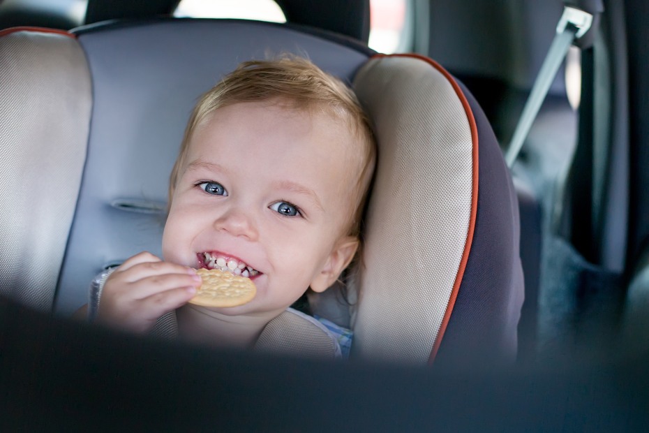 picture of a smiling toddler eating a cracker in the backseat of a car