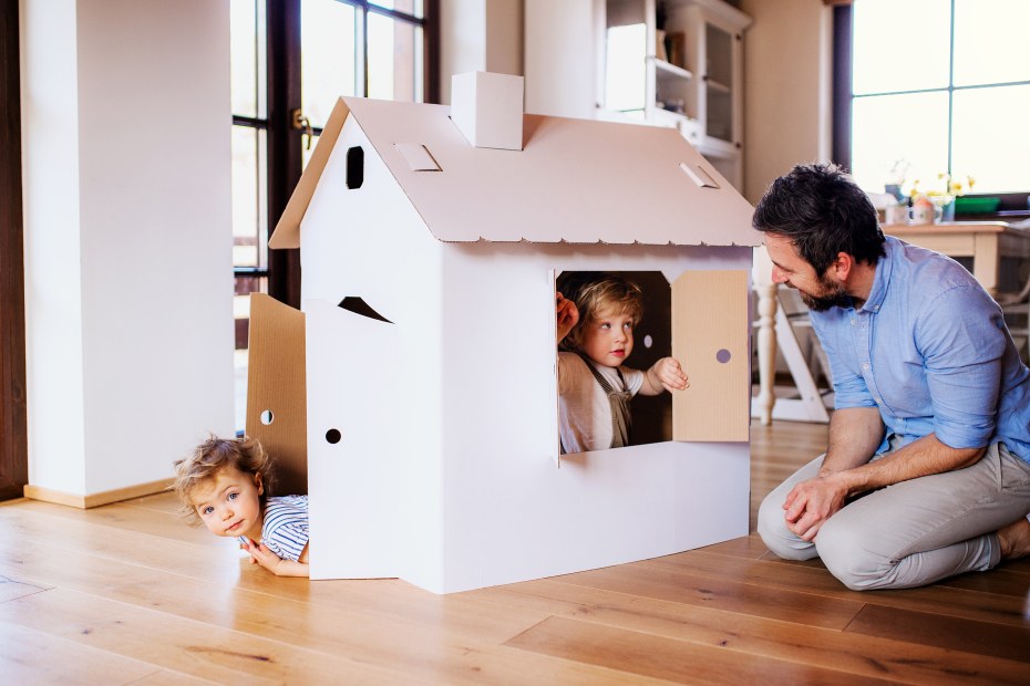 A father plays with toddlers inside a cardboard home.