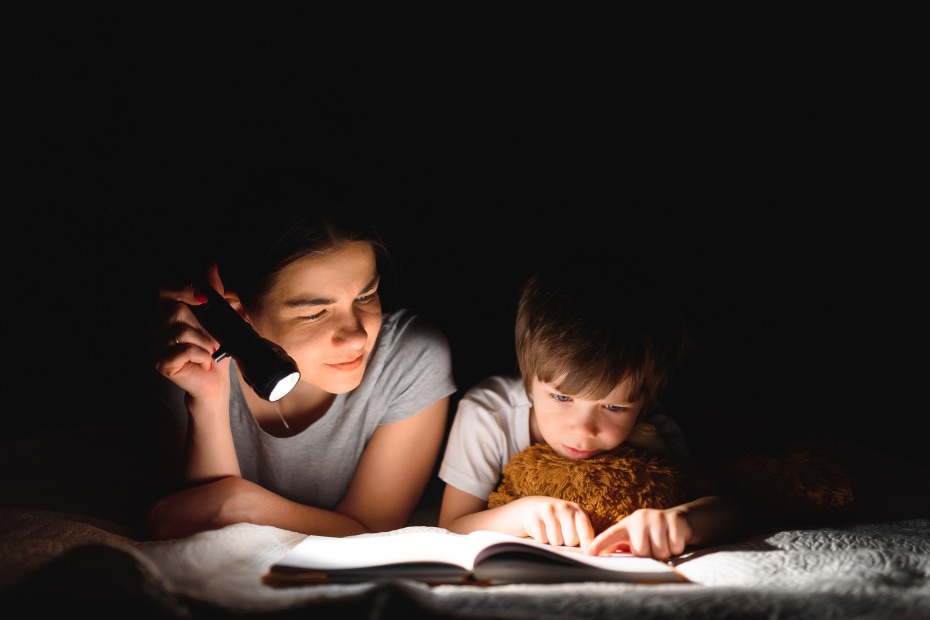 A mom reads a book with her child by flashlight.
