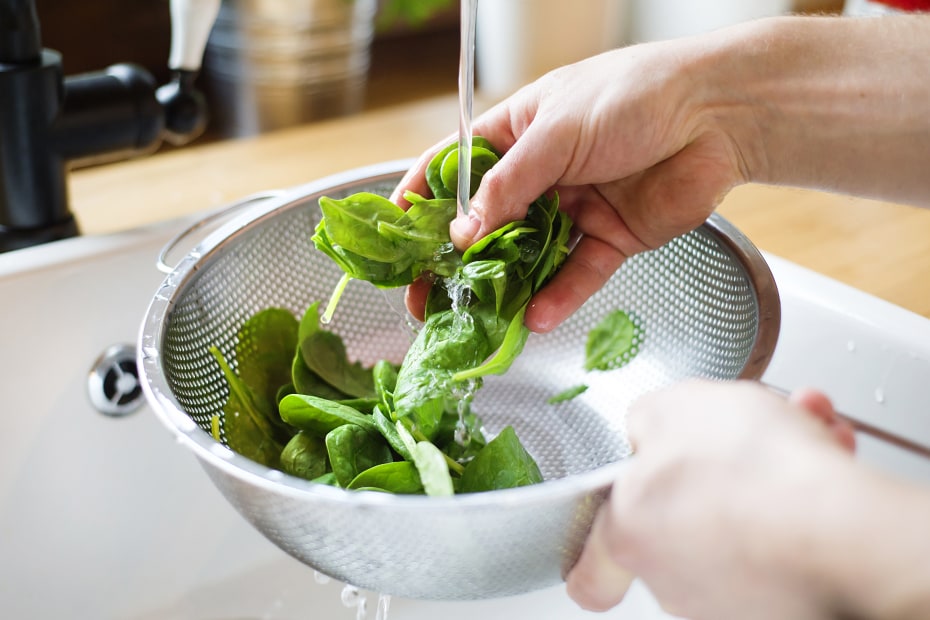 Spinach being washed in a collander under a running faucet.