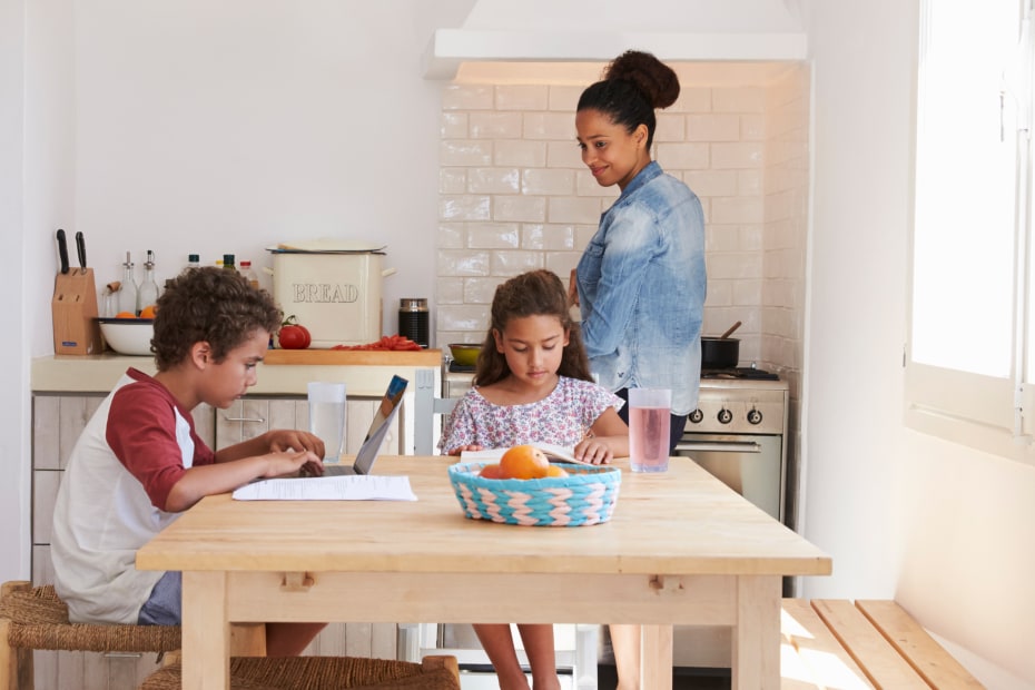 A mom cooks while her son and daughter do homework at the kitchen table.