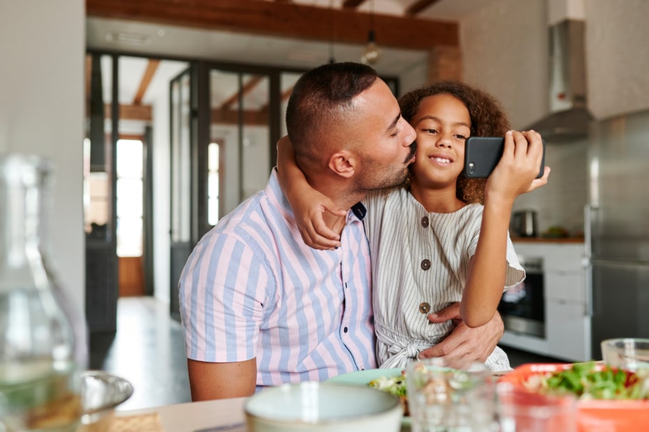 father kisses the cheek of his daughter while she looks at her phone in the family kitchen