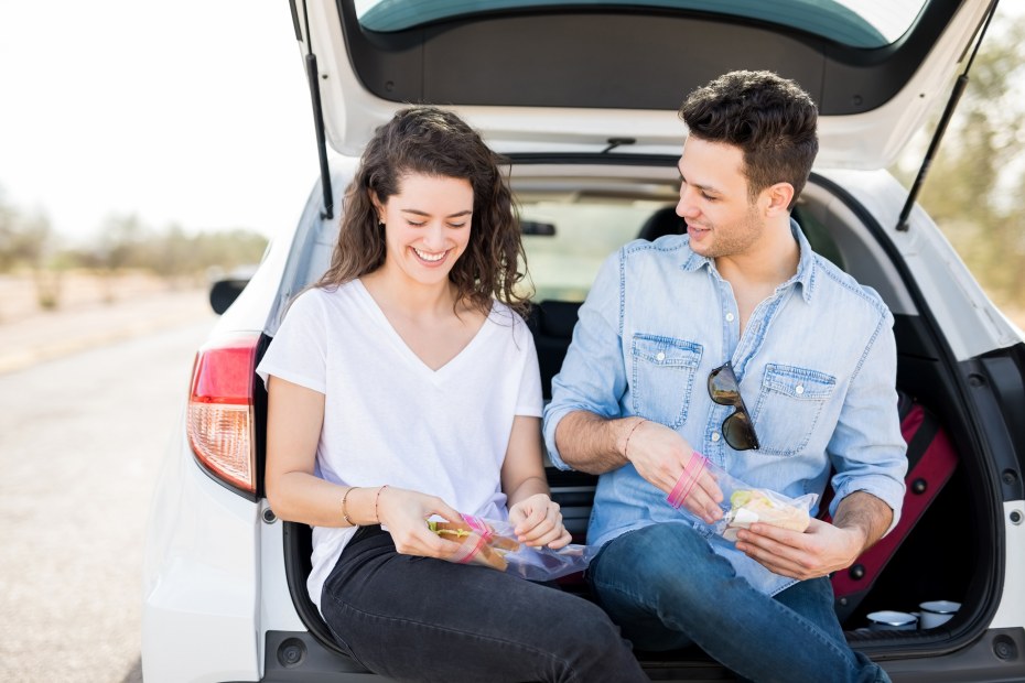 A couple sit on the tailgate and eat a sandwich on the side of the road.