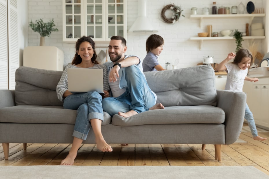 couple sits on gray couch looking at a laptop while their kids play in the background