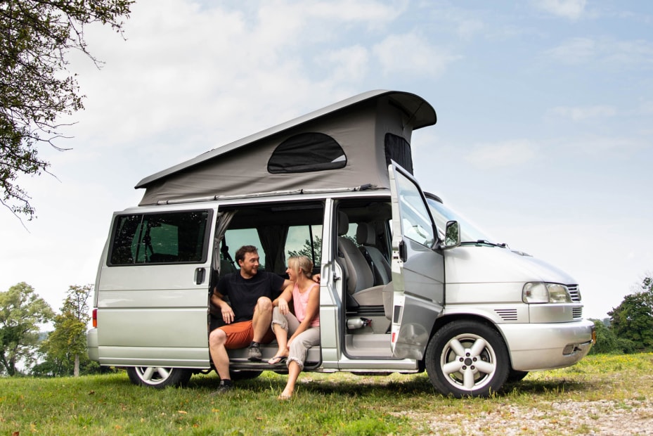 A couple sit in the open door of a class B camper van