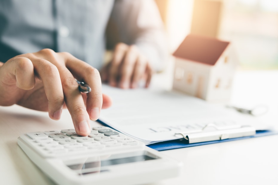 a hand holds a pen while typing on a calculator with paperwork and toy house in the background