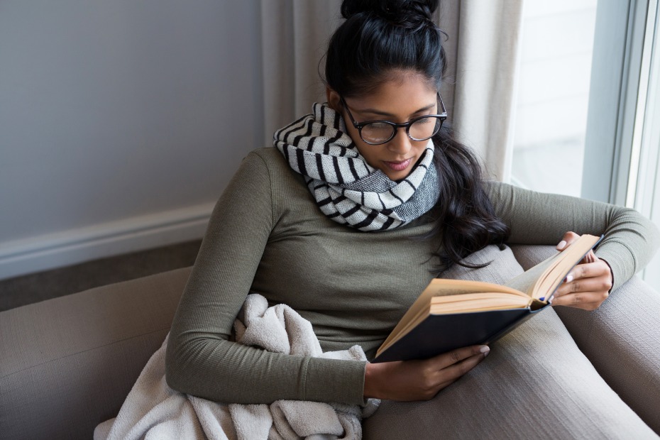 A woman wrapped in a blanket reads on the couch.