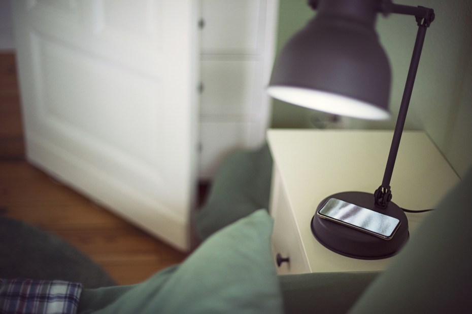 a black bedside lamp shines down on a charging smart phone in a bedroom with door ajar