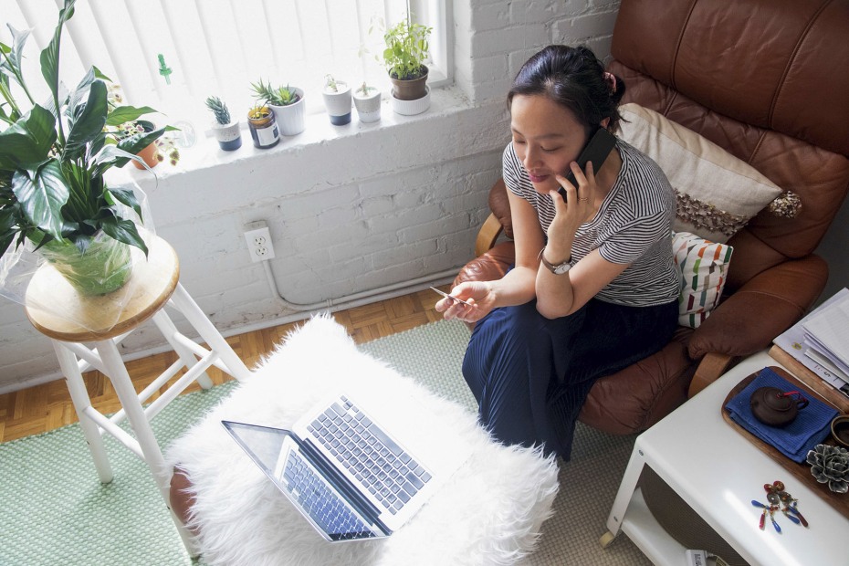 young woman on phone at home holds credit card while facing open laptop computer