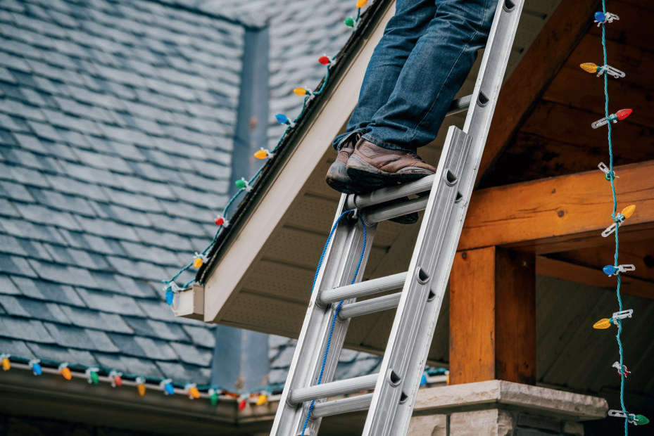 view of man from below the knees standing on ladder installing exterior holiday lights