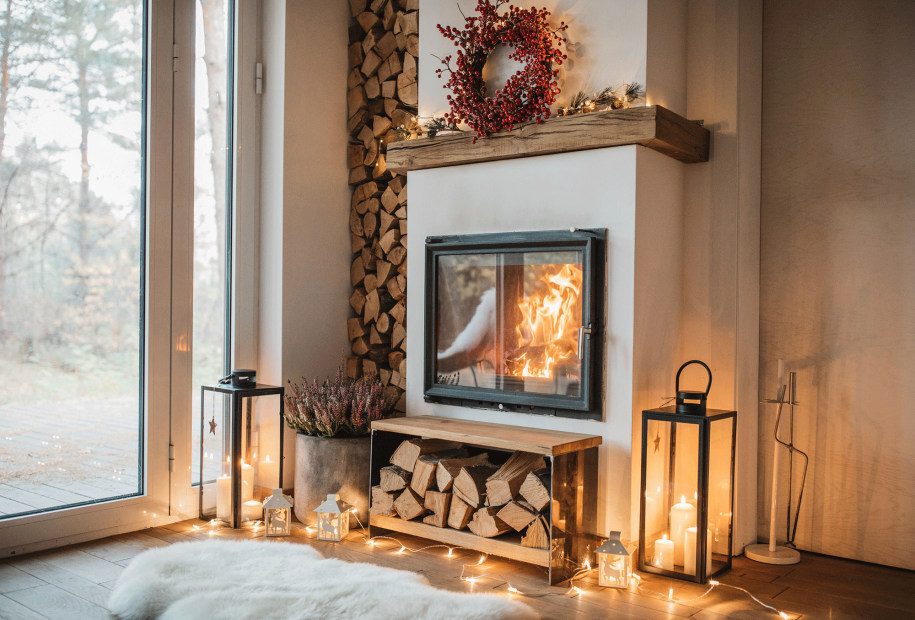 interior with active fireplace, split and stacked firewood, and holiday wreath hung on above on modern mantel beside framed glass door
