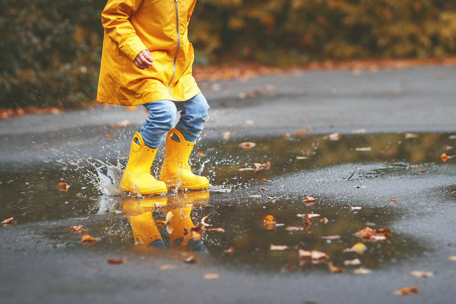 A young child in yellow rainboots and a yellow raincoat jumps in a big puddle.