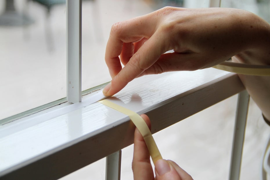 A woman uses weather stripping to seal gaps around a window.