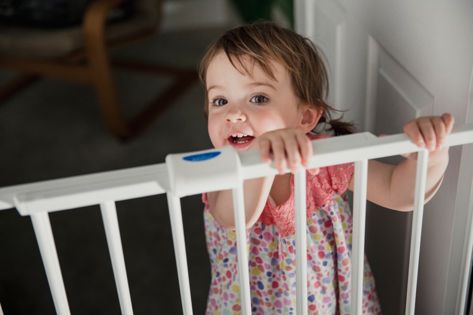 A little girl stands at a baby gate in a doorway.