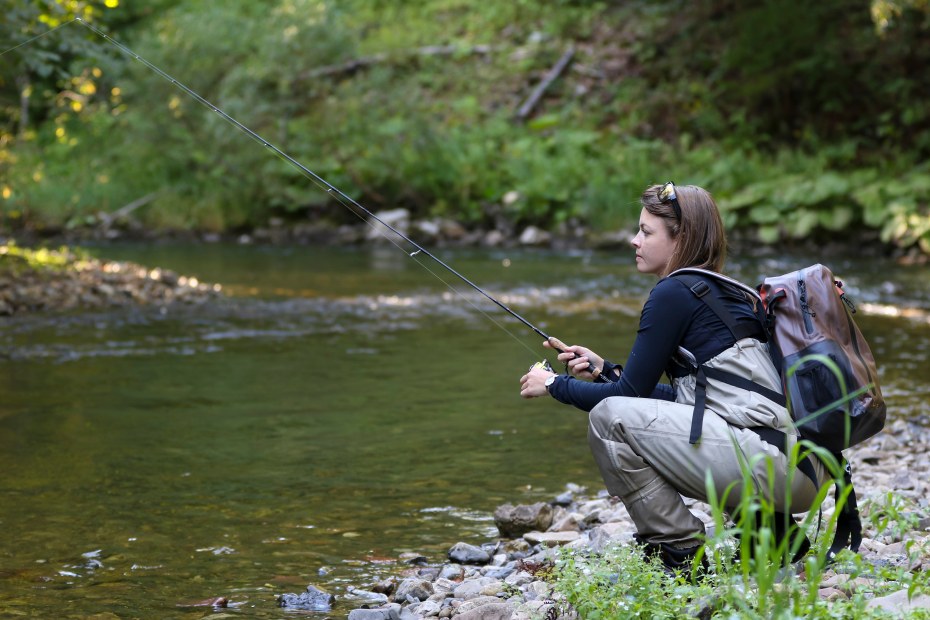 A woman fishes in waders alongside a river.