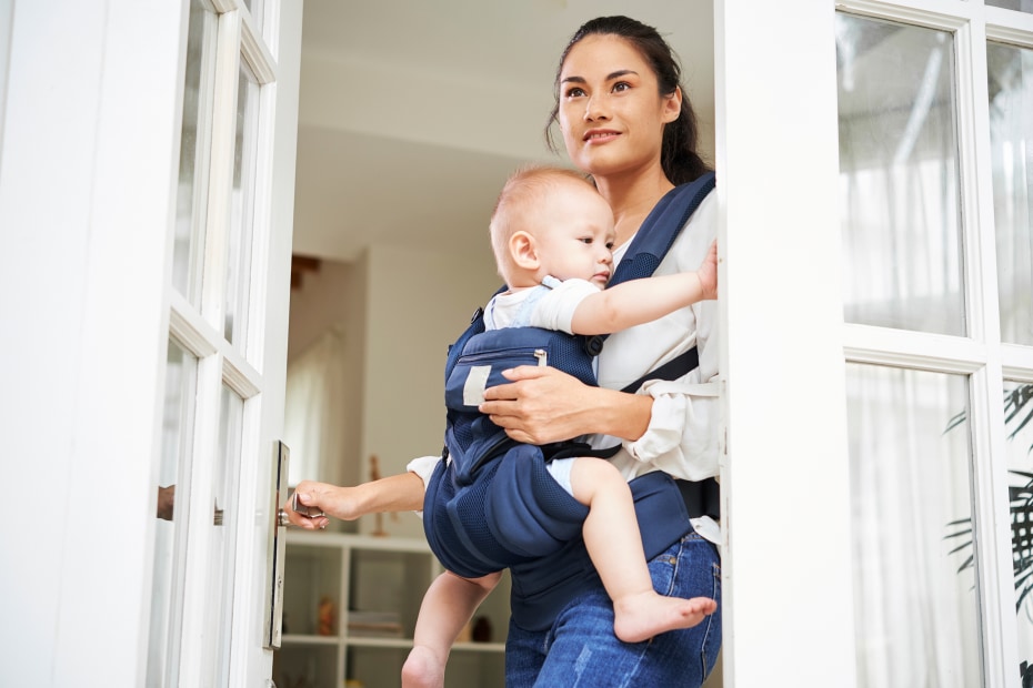 A woman with a baby in a carrier opens a French door.