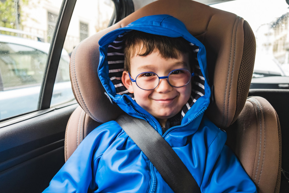 A boy in a bright blue raincoat and glasses sits in a tan booster seat in the backseat of a car.