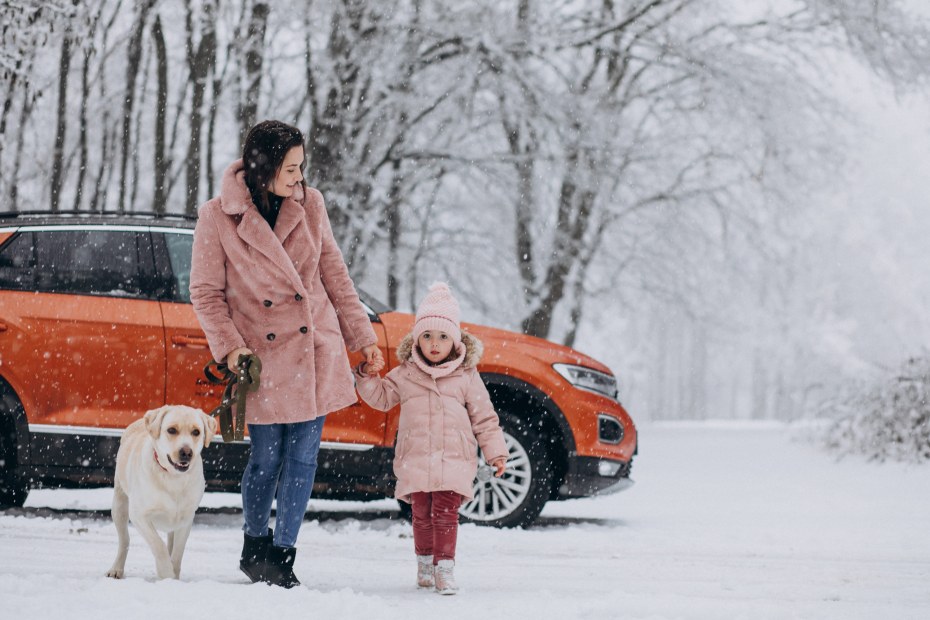 A woman and her daughter and her white labrador retriever walk down a snow covered street with an orange SUV in the background.