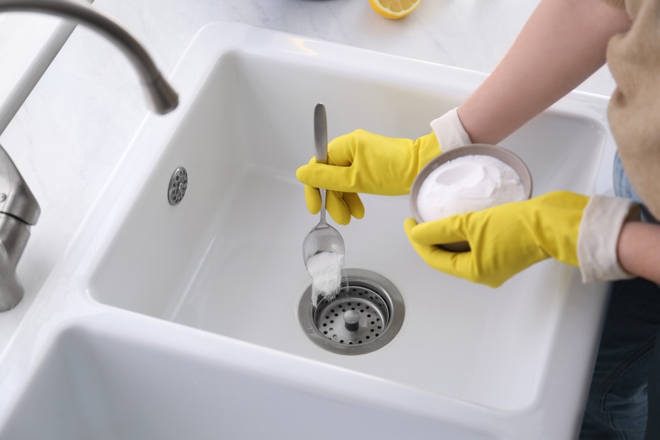 A person puts spoonfulls of baking soda into the kitchen sink to clean the drain pipe.