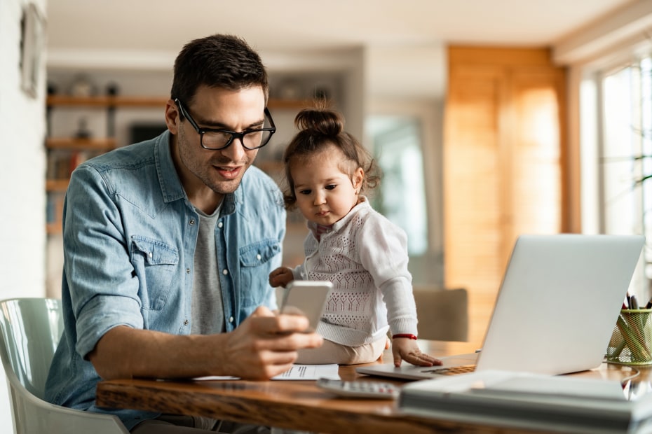 A dad sits with his daughter at the kitchen table while working on his phone.